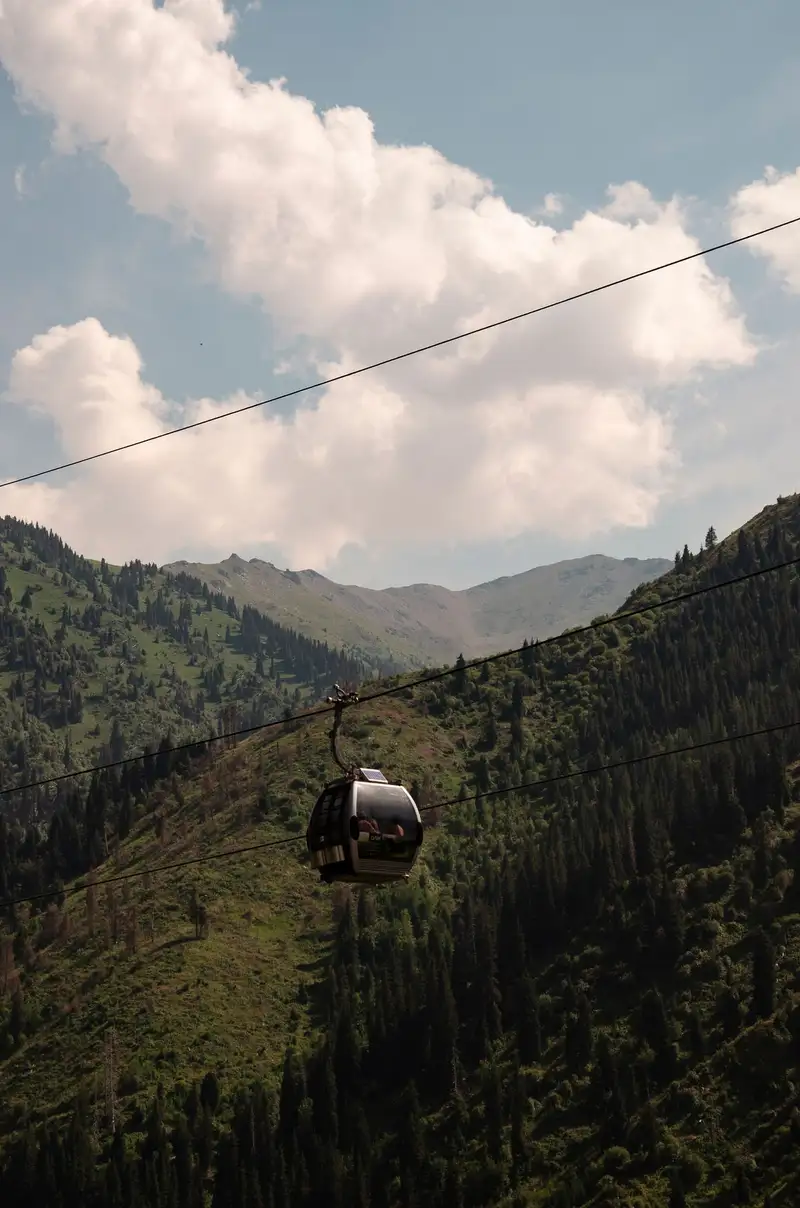 A cable-car gondola gliding above a green, spruce-covered mountain gorge under summer clouds